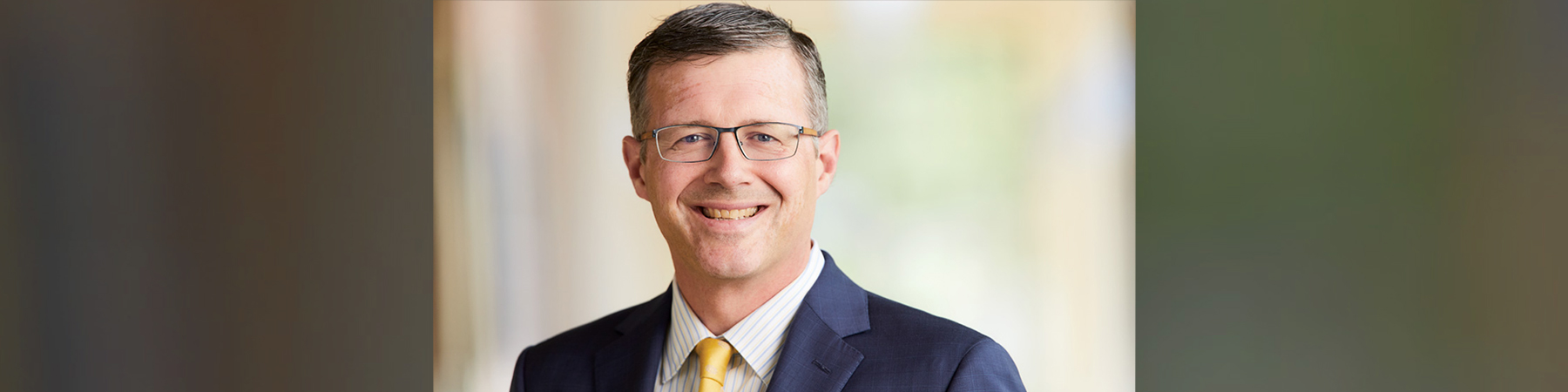 Provost, Douglas Woods, who wears glasses, is smiling and wearing a dark navy blue suit, white shirt, and a gold tie.