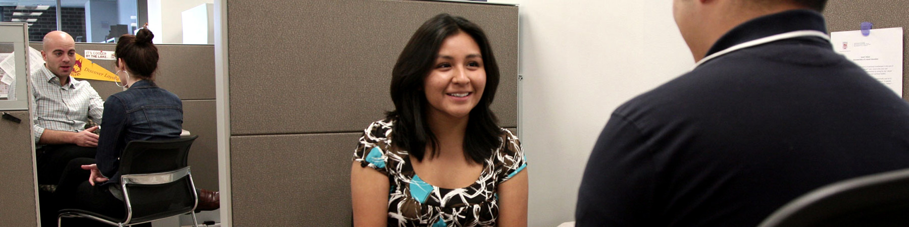 Two students are seated on opposing cubicles meeting with their academic advisors.