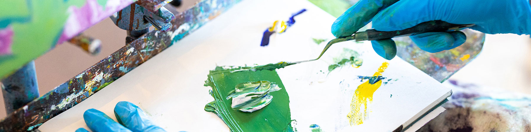 An unseen student mixes green paint on a palette with a palette knife.