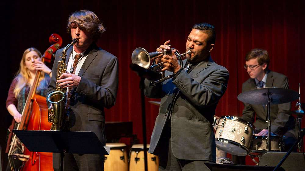 A small group of musicians play music. One saxophonist and one trumpeter, both males, stand out front.