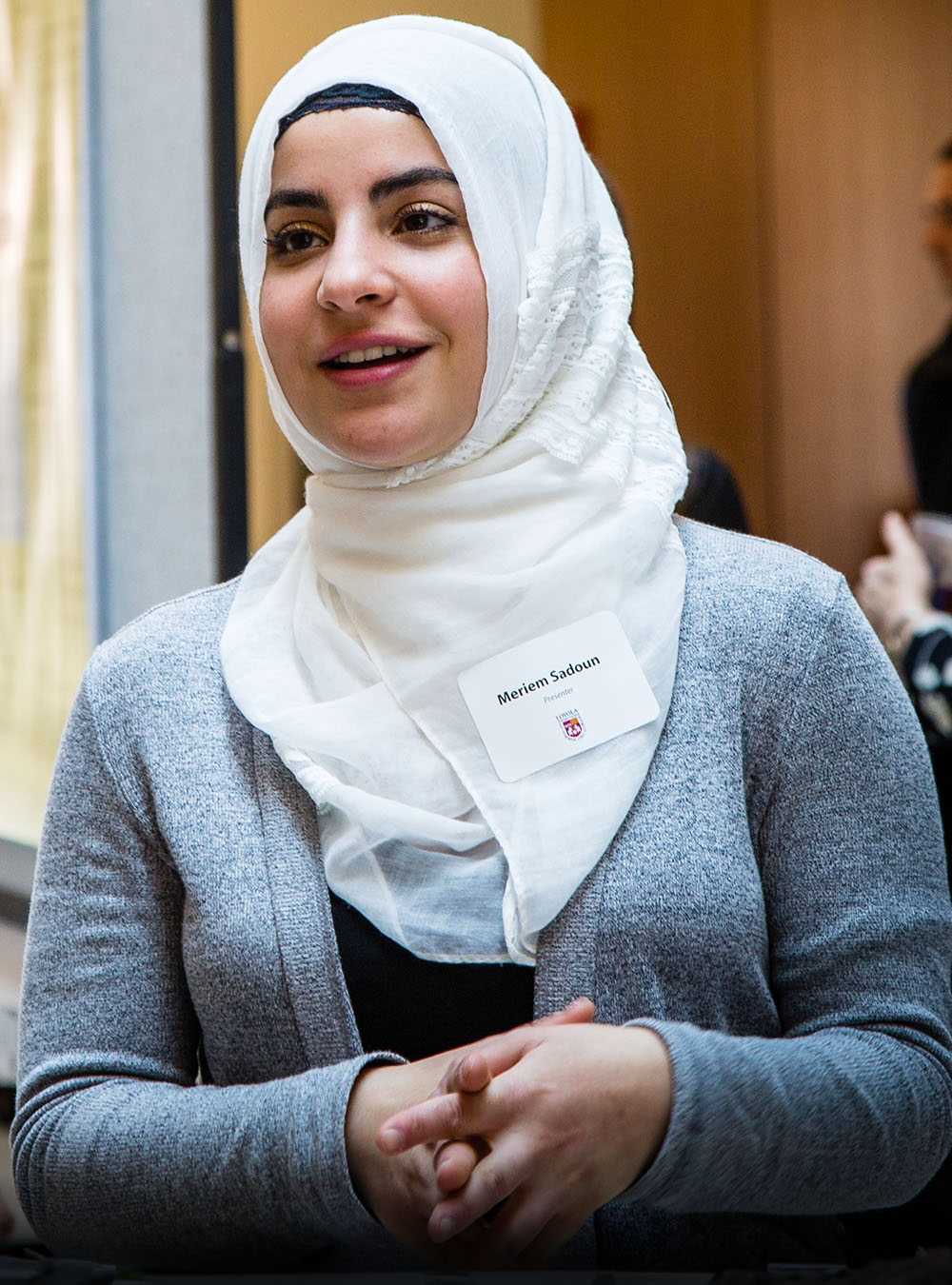 A woman in a white hijab and gray sweater stands indoors, wearing a name tag. She appears to be speaking or engaged in conversation.