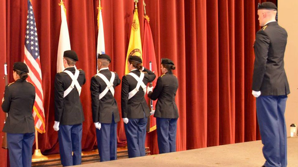 A color guard consisting of five uniformed individuals in navy blue pants, black jackets, and white cross belts presents flags on a stage with a red curtain backdrop. Another uniformed individual stands to the right, observing.