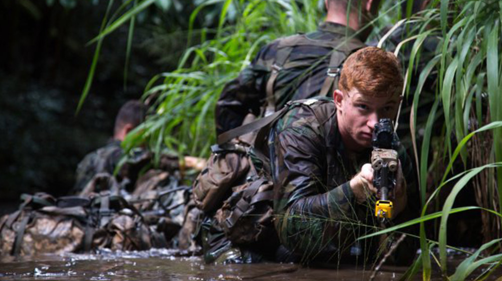 A military trainee is wading through a jungle river holding an assault rifle with a yellow BFA attachment.