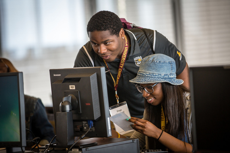 Two African American Loyola University Chicago students work together at a computer screen. The male is wearing a Loyola lanyard around his neck, standing and leaning over the female, wearing a blue bucket hat, looking at the computer screen.