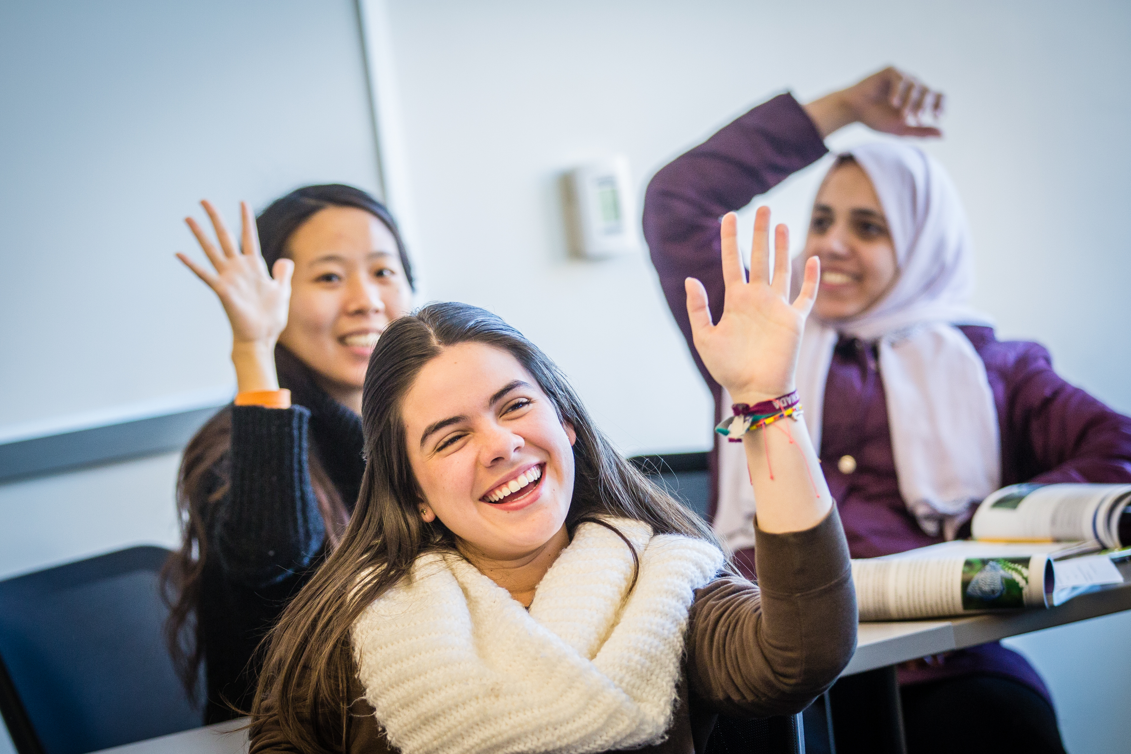 Group of three diverse international, female, Loyola University Chicago students sitting at desks in a classroom. They have their hands raised and are laughing and smiling.