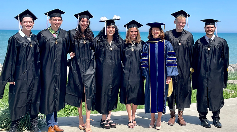 A small group of graduates dressed in caps and gowns stand shoulder to shoulder with Lake Michigan as a backdrop.