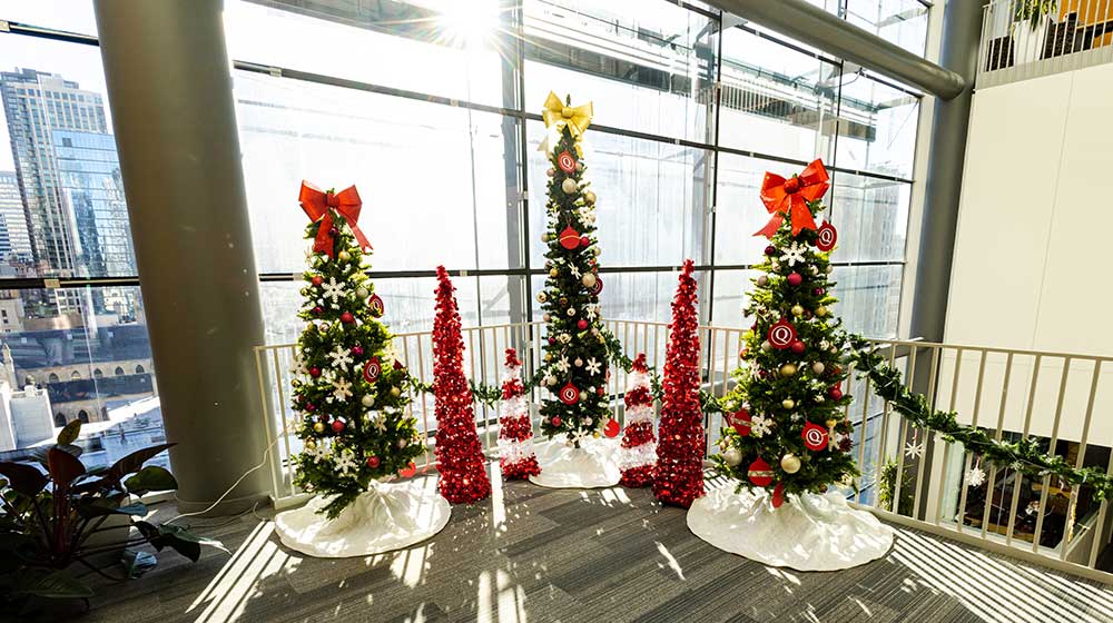 Three green Christmas trees and two smaller red trees stand in the Schreiber Center in front of the Chicago skyline.
