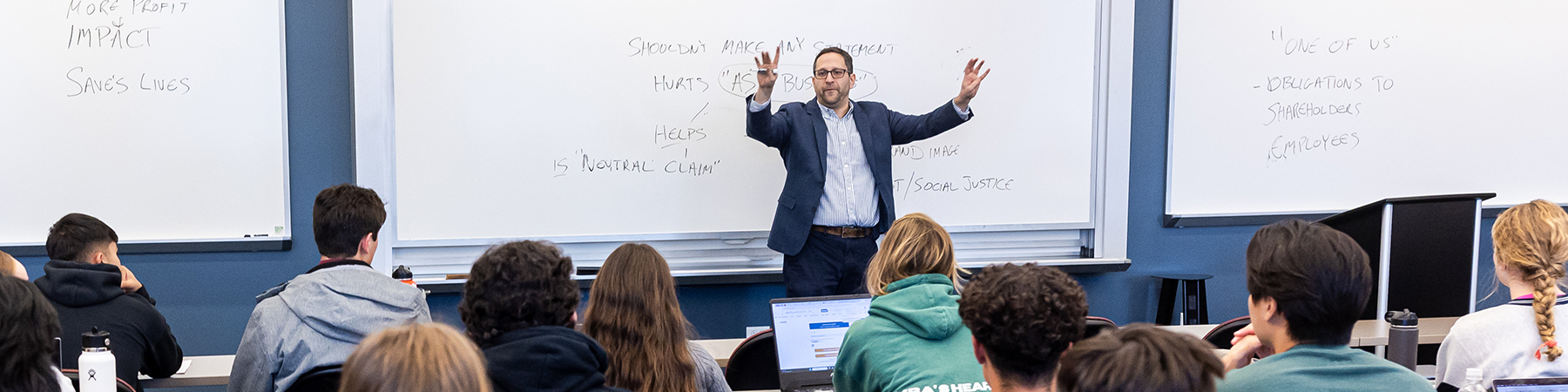 Quinlan professor Abe Singer, with his hands raised out toward a classroom of engaged students, intructs them as he stands in front of a white board at the front of the room.