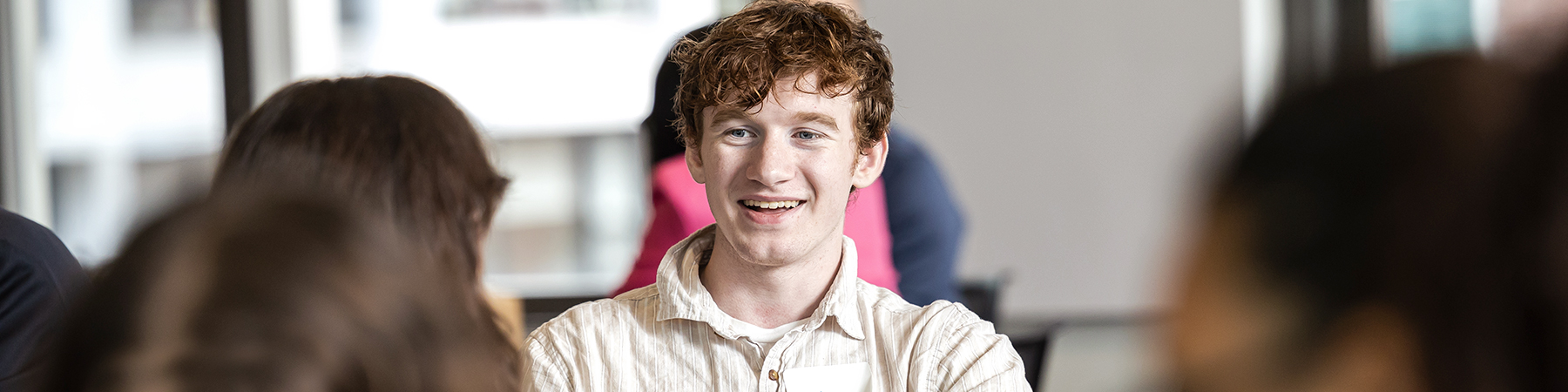 A smiling young man with red curly hair sits with other student at a table. He is facing the camera and engaged in conversation. He wears a light short sleeved, collared shirt.