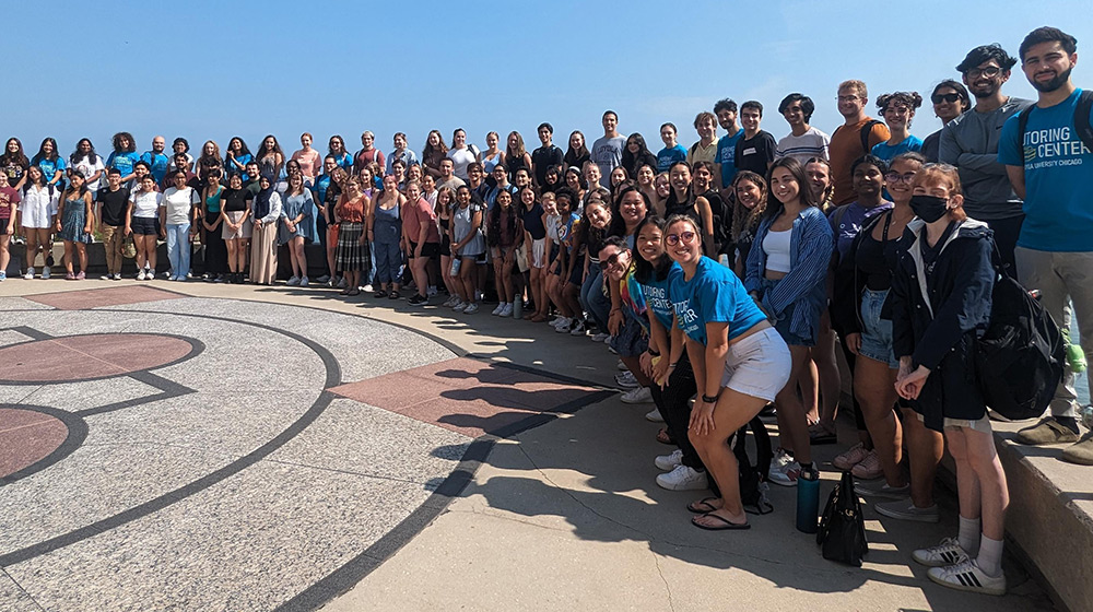 A large gathering of dozens of students pose along the lakeshore for a group photo. They are dressed in casual attire and Loyola University Chicago apparel.