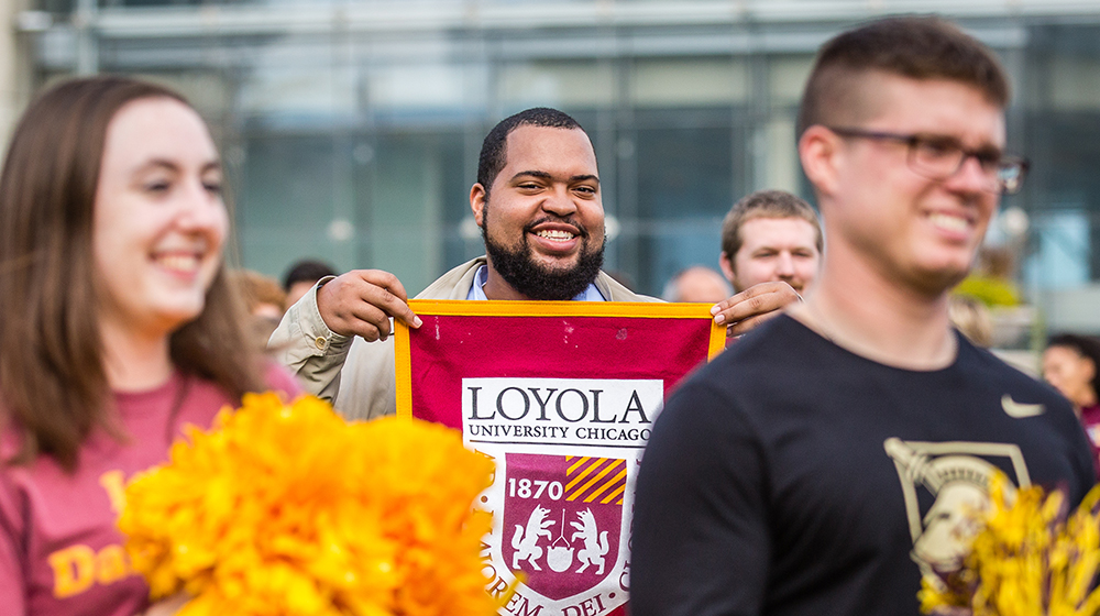 A student in focus is smiling directly at the camera and holding a chest-width flag with the Loyola University Chicago crest on it. On either side are students out of focus holding pom-poms while looking at something off screen.